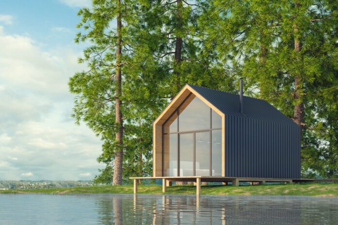 Ein Tiny House steht auf einem Steg direkt an einem weitläufigen See in einer Waldlichtung.