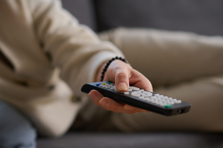 Eine Frau liegt auf dem Sofa und schaltet mit der Fernbedienung den Fernseher um.