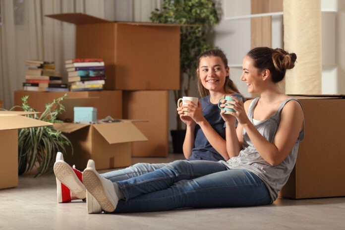 Zwei Frauen sitzen mit Tassen in der Hand vor Umzugskisten in der Wohnung. Sie gründen wohl eine WG.